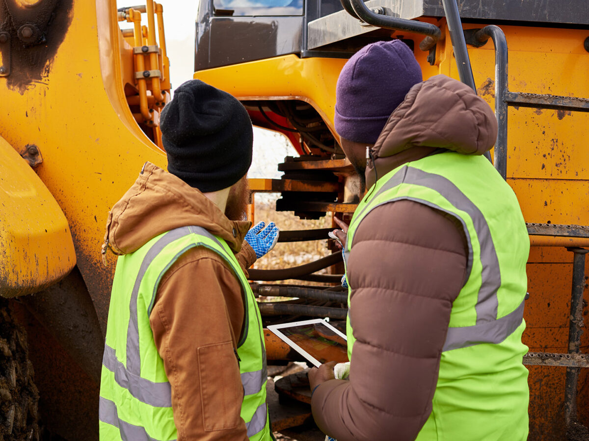 2 male construction workers examining a piece of construction equipment while holding an electronic tablet.