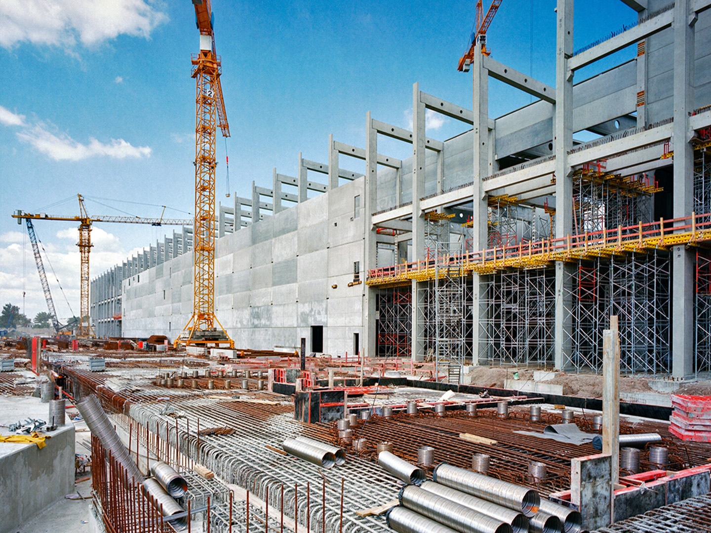 A large scale industrial construction site with 3 cranes, piles of piping and steel rebar.